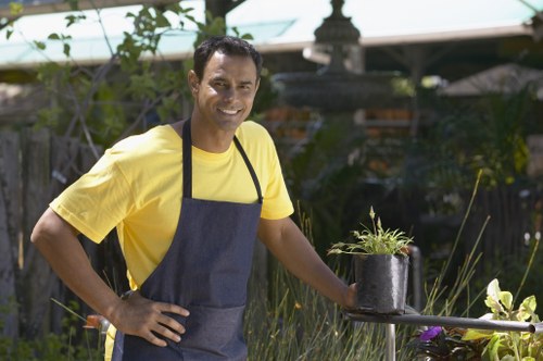 A smiling man wearing a yellow T-shirt and a dark apron stands in a well-maintained garden in Thornton Heath, surrounded by lush green plants, shrubs, and a mix of grass and garden borders. He is holding a black watering can with a small potted plant on a metal table nearby. The garden features a paved area, some decorative elements, and a wooden trellis or fencing in the background, with mature trees and flowering plants contributing to a vibrant outdoor space. The scene is illuminated by natural daylight, suggesting a mild weather day. This image reflects professional gardening and outdoor maintenance services, including plant care and garden landscaping, relevant to companies like Lawn Mowing Thornton Heath operating in the local area.